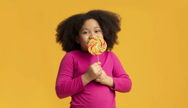 Olumlu küçük afro kız lolipop yalıyor, şekerden zevk alıyor. — Stok fotoğraf