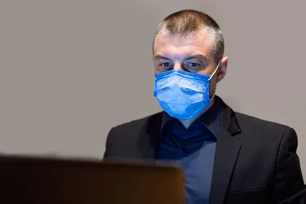 Biznesmen pracujący na laptopie nosząc maskę medyczną siedzi, szary tle — Zdjęcie stockowe