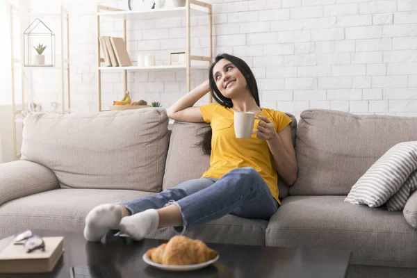 Blijf thuis, drink koffie. Ontspannen Spaanse meisje genieten van haar ochtend koffie op de bank in de woonkamer — Stockfoto