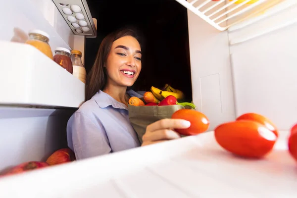 Cheerful woman unpacking food bag into fridge