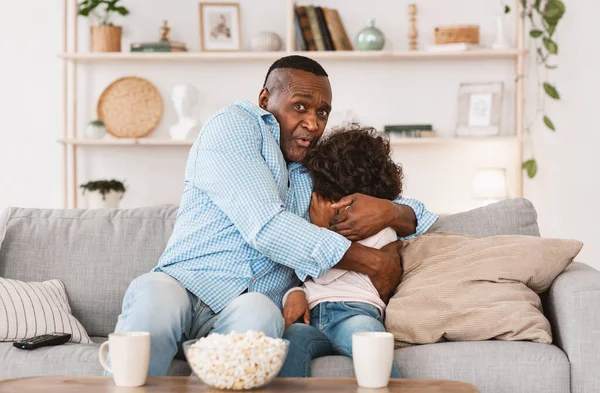 Családi mulatságok. Afrikai-amerikai nagypapa ölelgeti rémült unokáját, miközben otthon filmet néz. — Stock Fotó