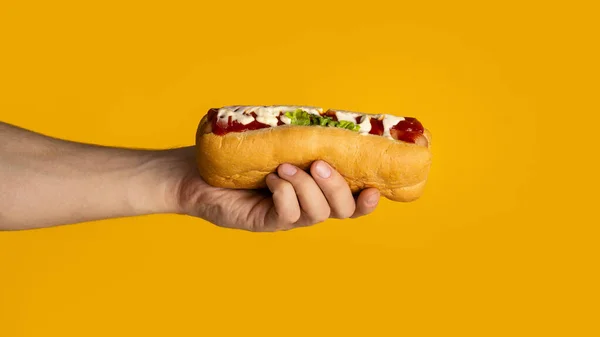 Primo piano del giovane che tiene fresco hot dog con salsa su sfondo arancione, panorama — Foto Stock