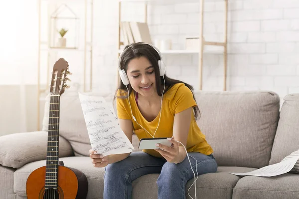 Stay home, stay creative. Joyful girl in headphones with music notes watching acoustic guitar tutorial on mobile phone