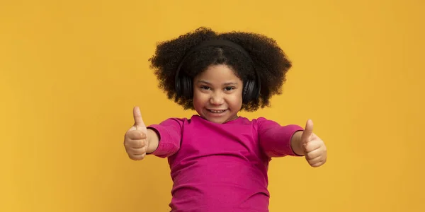 Niña positiva escuchando música en auriculares inalámbricos y mostrando el pulgar hacia arriba —  Fotos de Stock