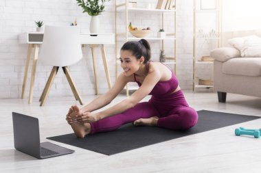 Kendini izole etme egzersizi. Spor giyim sektöründe neşeli bir kız yoga yapıyor ya da evde online öğretmenlik yapıyor.