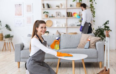 Ev işi. Kadın masayı siliyor, erkek tozu siliyor.