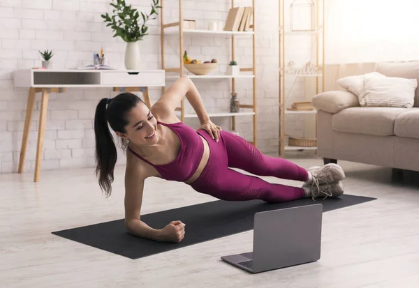 코로나 바이러스 격리 구역. Fit woman doing side plank while watching online sports tutorial at home — 스톡 사진