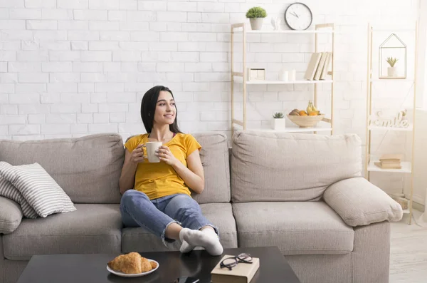 Fica em casa, bebe café. Menina milenar com xícara de bebida quente relaxante no sofá confortável na sala de estar, espaço de cópia — Fotografia de Stock