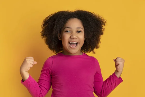 Gioiosa bambina nera che celebra il successo con pugni alzati, sfondo giallo — Foto Stock
