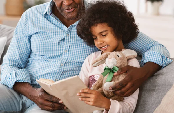 Reading together. African American girl with bunny and her granddad with fascinating book at home