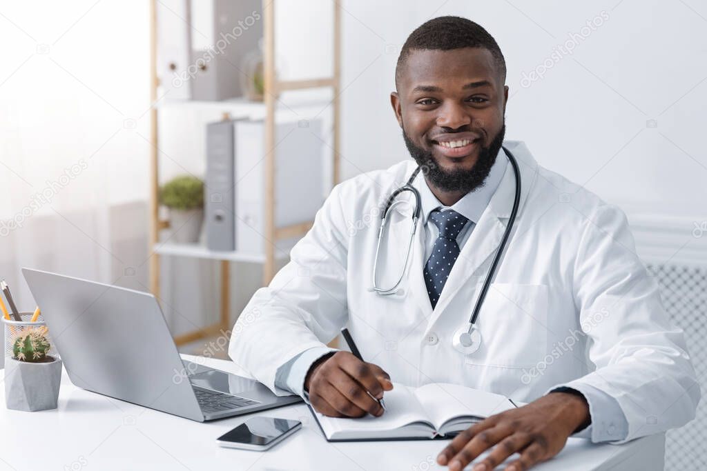 Happy black physician taking notes while working online