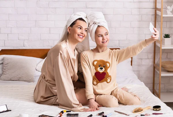 Spa di famiglia a casa. Felice mamma e figlia con cosmetici e asciugamani da bagno prendendo selfie in camera da letto — Foto Stock
