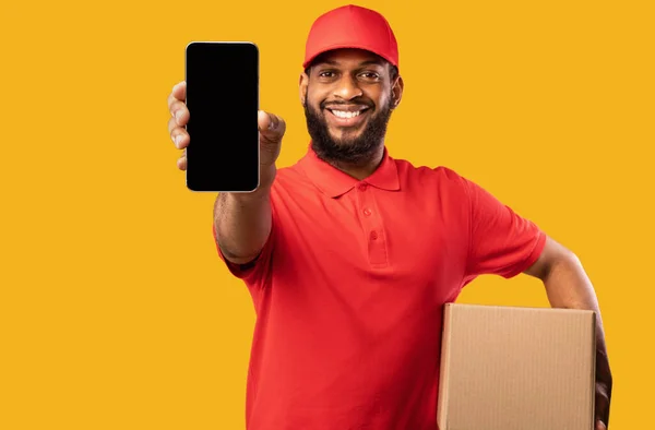 Africano Courier mostrando Smartphone branco tela segurando caixa, fundo amarelo — Fotografia de Stock