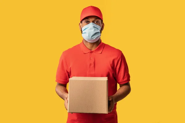 Preto Courier cara entregando caixa de papelão vestindo máscara, fundo amarelo — Fotografia de Stock