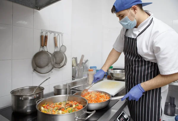 Young man cooking food for delivery in mask and gloves
