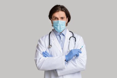 Koruyucu tıbbi maskeli, kendine güvenen erkek doktor elinde aşı şırıngası tutuyor.