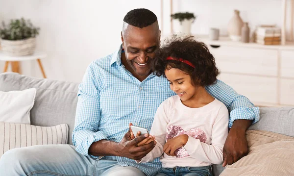 Gammal afroamerikansk man och hans barnbarn använder smartphone, spela TV-spel eller surfa på webben — Stockfoto