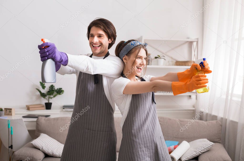 Funny young couple fooling while cleaning apartment, aiming with detergent sprayers