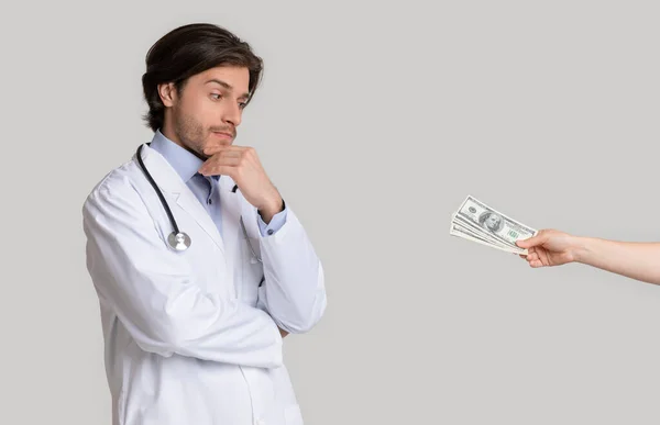 Сумнівний молодий лікар дивиться на грошовий хабар, запропонований пацієнткою — стокове фото