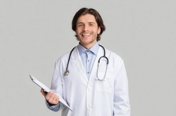 Mosolygó fiatal férfi orvos fehér köpenyben sztetoszkóppal és írótáblával — Stock Fotó