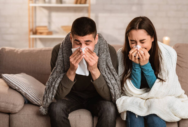 Sick young couple blowing noses sitting on couch, having flu