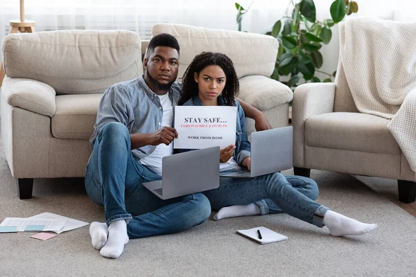 Sério casal africano sentado com laptops no chão e segurando cartaz de aviso — Fotografia de Stock