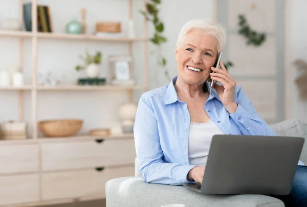 Сучасна старша жінка використовує ноутбук і розмовляє на мобільному телефоні вдома — стокове фото