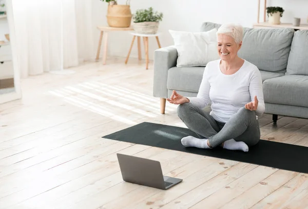 Joga online. Zrelaksowana seniorka medytująca przed laptopem w domu — Zdjęcie stockowe