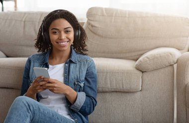 Gülümseyen Afrikalı kadın kulaklık takıyor, evde akıllı telefondan müzik dinliyor.