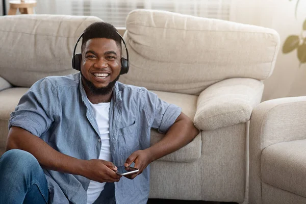 Fröhlicher Schwarzer mit Kopfhörer, der zu Hause Musik auf dem Smartphone hört — Stockfoto