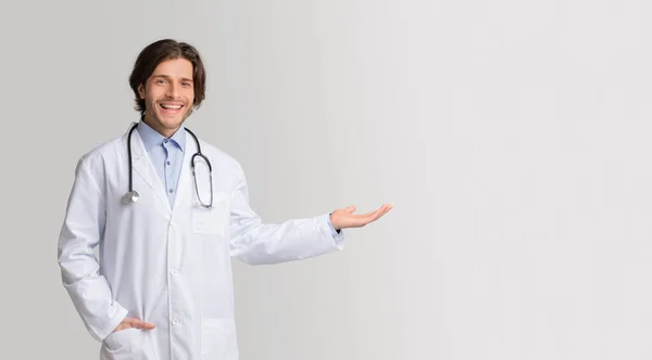 Κλινική διαφήμιση. Φιλικός νεαρός γιατρός δείχνοντας προς τα έξω στο χώρο αντίγραφο με το χέρι — Φωτογραφία Αρχείου