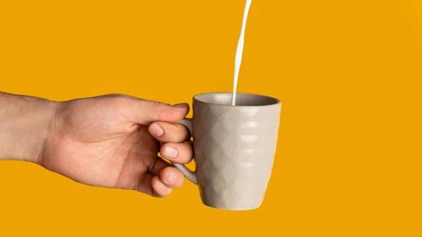 Joven irreconocible sosteniendo una taza de bebida caliente con leche vertiendo en ella, fondo naranja. Panorama — Foto de Stock