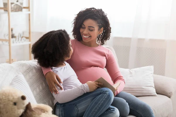 Genç hamile siyah kadın evde küçük kızıyla kitap okuyor. — Stok fotoğraf