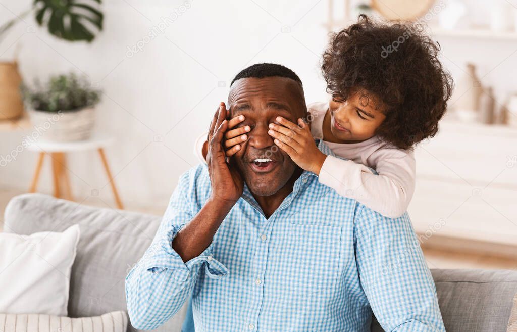 African American girl closing her granddads eyes from behind, surprising him or playing guess who game