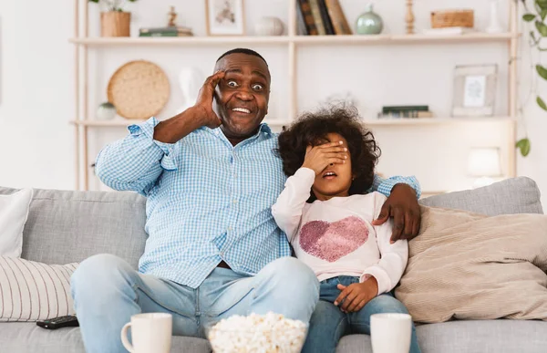 A családi mulatságok lezárása. Megdöbbent afro-amerikai férfi és rémült unoka néz horror film otthon — Stock Fotó