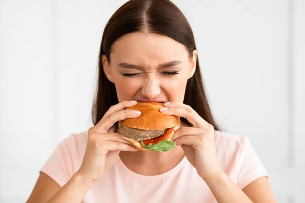 Menina mordendo hambúrguer desfrutando de junk food ter fraude refeição interior — Fotografia de Stock