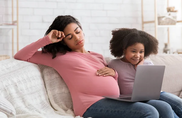 Cansada mujer embarazada sintiéndose agotada mientras usa el ordenador portátil con su pequeña hija — Foto de Stock