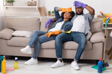 Finally Done. Exhausted black couple sitting on couch after spring-cleaning house clipart
