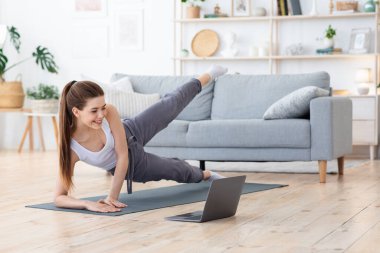 Spor yapan, evde egzersiz yapan, dizüstü bilgisayarda video izleyen neşeli bir kadın.