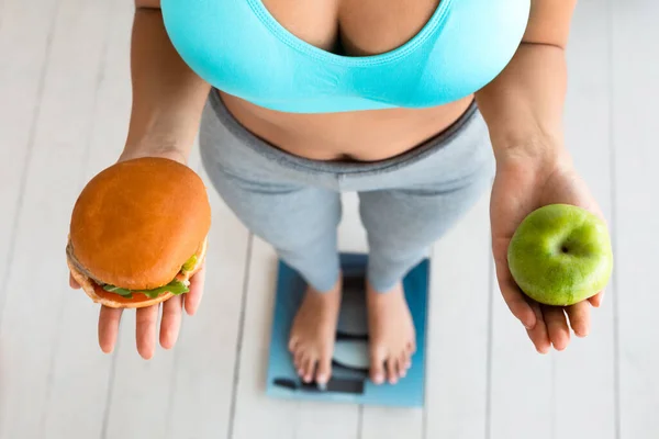 Mulher escolhendo entre hambúrguer e maçã em pé em balanças de peso, cortado — Fotografia de Stock