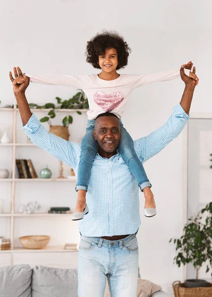 Jogos engraçados com o avô. Homem afro-americano sênior carregando sua neta nos ombros em casa — Fotografia de Stock