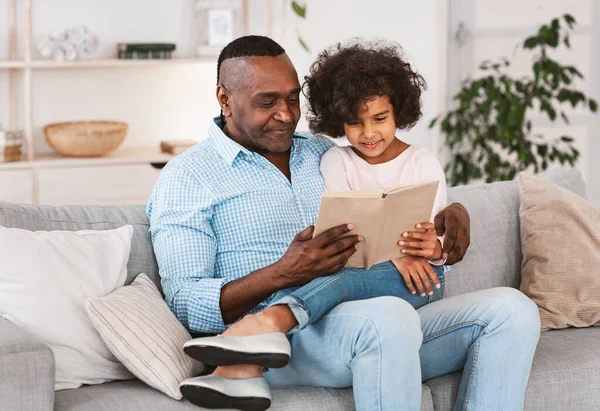 Interessante ficar passatempos em casa. Menina muito afro-americana ouvindo o avô ler livro na sala de estar — Fotografia de Stock