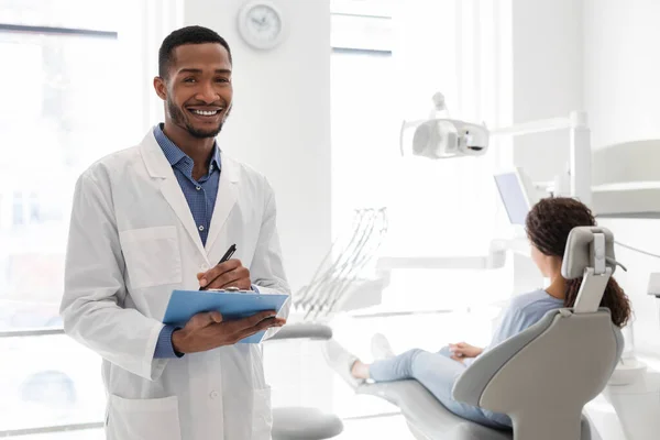 Odontólogo negro sonriendo y sosteniendo la carpeta en el gabinete dental — Foto de Stock