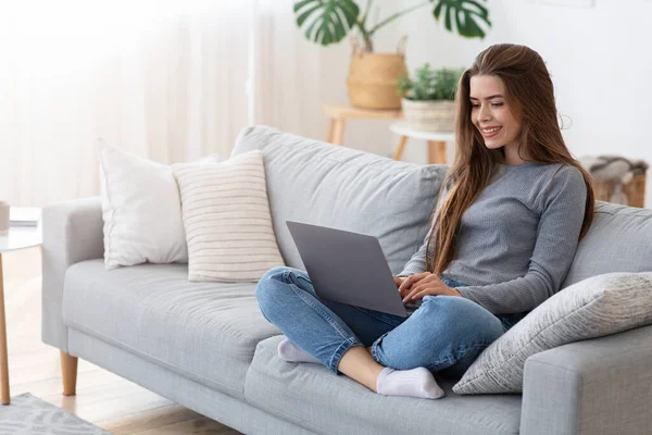 Sorridente giovane signora seduta con computer portatile sul divano a casa — Foto Stock