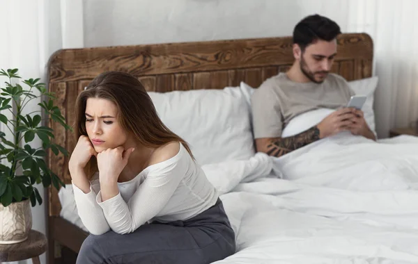 Ofendido menina sentado na cama, homem olha para o telefone — Fotografia de Stock