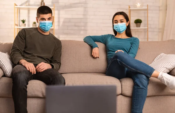 Casal em máscaras usando laptop sentado no sofá — Fotografia de Stock