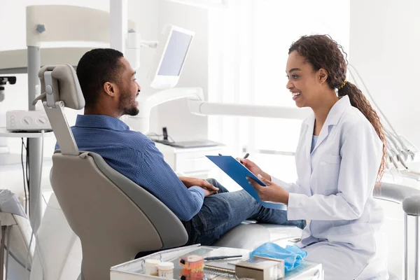 Dentista sorridente preenchendo prontuário médico, conversando com paciente — Fotografia de Stock