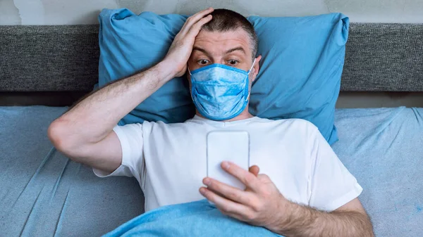 Szokujący człowiek w medycznym masce ochronnej przy użyciu smartfona w łóżku — Zdjęcie stockowe