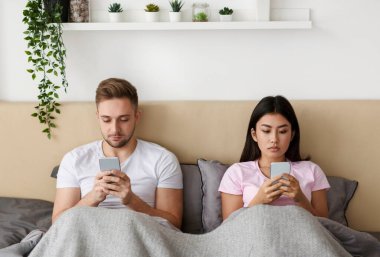 Çift akıllı telefon kullanıyor. Birbirlerini içeri tıkıyorlar. Yatakta oturuyorlar.