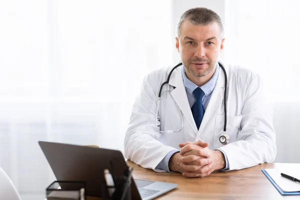 Portret dojrzałego lekarza patrzącego na kamerę siedzącą przy biurku — Zdjęcie stockowe
